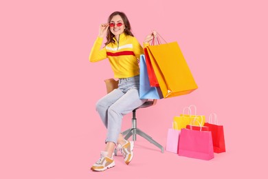 Photo of Happy woman in stylish sunglasses holding many colorful shopping bags on armchair against pink background