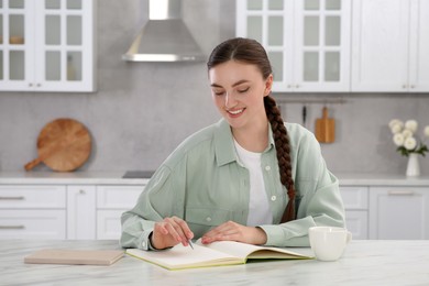 Photo of Beautiful young woman writing in notebook at white marble table indoors