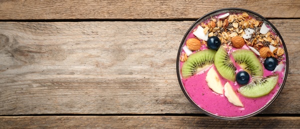 Photo of Delicious acai smoothie with granola and fruits in dessert bowl on wooden table, top view. Space for text