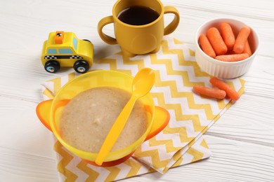 Photo of Baby food. Puree in bowl, small carrots, toy and drink on white wooden table