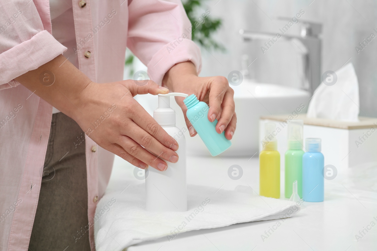 Photo of Woman pouring cosmetic product into plastic bottle at white countertop in bathroom, closeup. Bath accessories