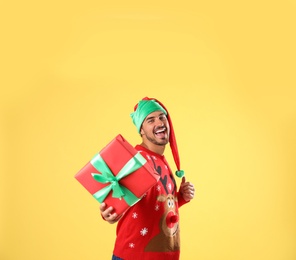 Photo of Happy man in Christmas sweater and elf hat holding gift box on yellow background