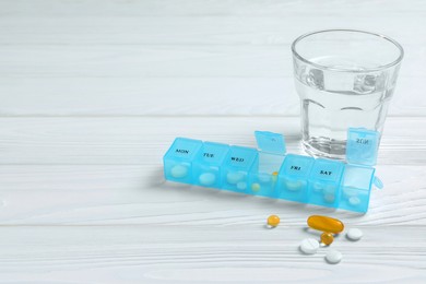 Photo of Weekly pill box with medicaments and glass of water on white wooden table. Space for text