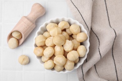 Photo of Bowl and scoop with peeled Macadamia nuts on white tiled table, flat lay