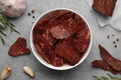 Photo of Delicious beef jerky in bowl and seasoning on light gray table, flat lay