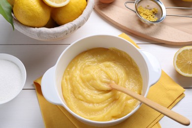 Photo of Delicious lemon curd in bowl, ingredients, spoon and sieve on white wooden table, above view