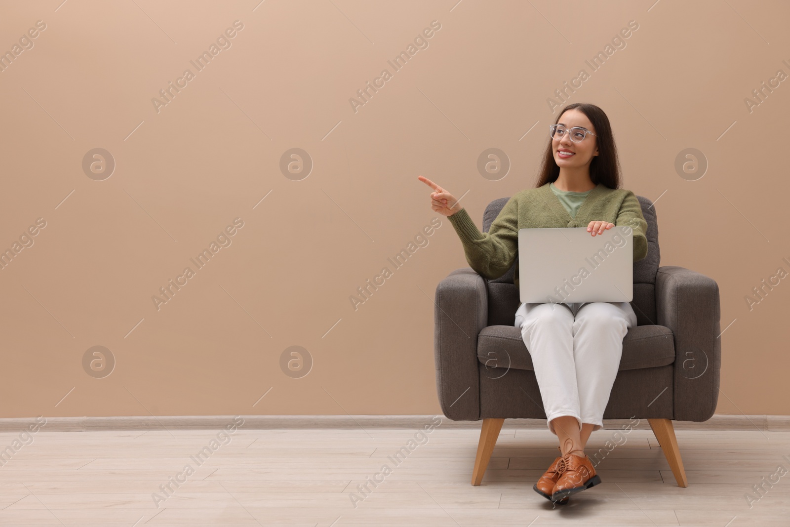 Photo of Beautiful woman with laptop pointing on something while sitting in armchair near beige wall indoors, space for text