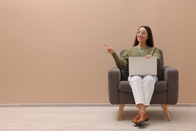 Photo of Beautiful woman with laptop pointing on something while sitting in armchair near beige wall indoors, space for text