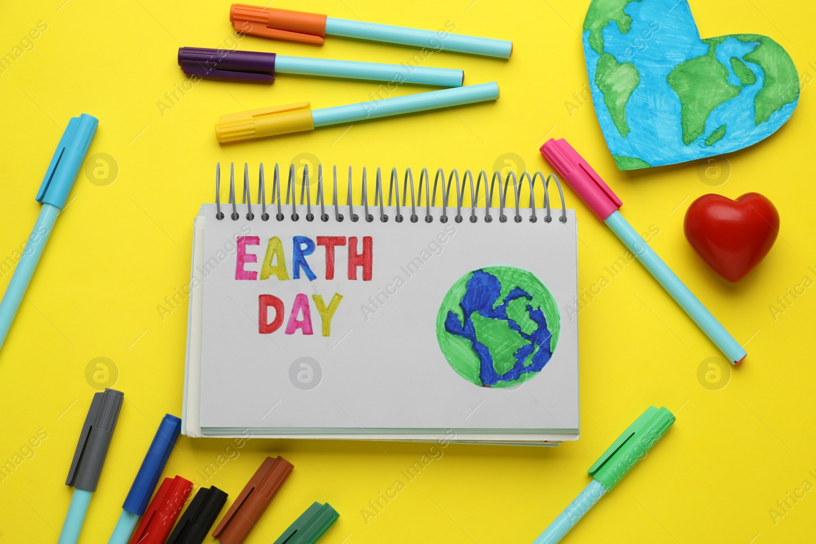 Photo of Notebook with phrase Earth Day, heart shaped model of planet and felt pens on yellow background, flat lay