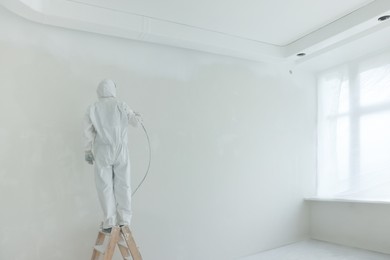 Photo of Decorator painting wall on ladder near windows in room, back view