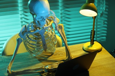 Photo of Waiting concept. Human skeleton sitting at wooden table with laptop indoors