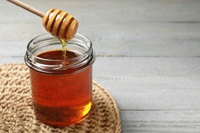 Pouring tasty honey from dipper into glass jar at grey wooden table, closeup. Space for text