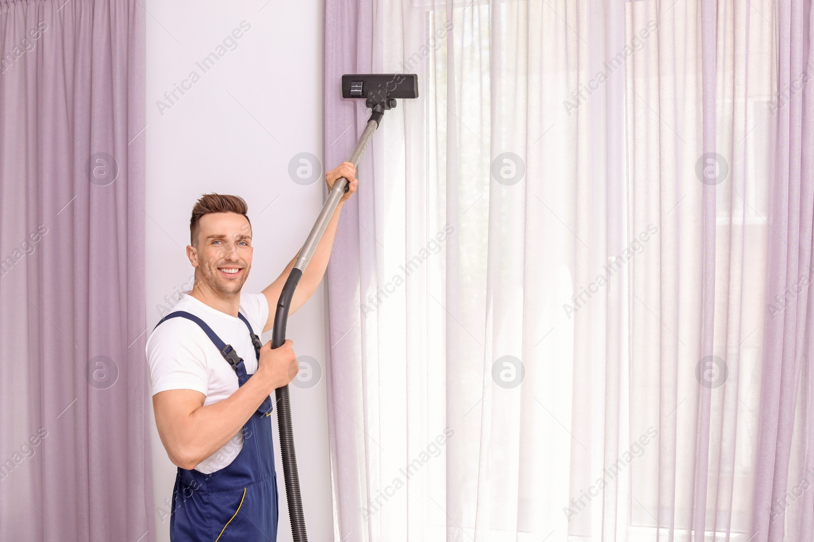 Photo of Male worker removing dust from curtains with professional vacuum cleaner indoors