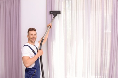 Male worker removing dust from curtains with professional vacuum cleaner indoors