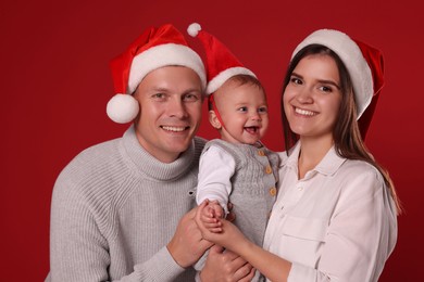 Happy couple with cute baby wearing Santa hats on red background. Christmas season