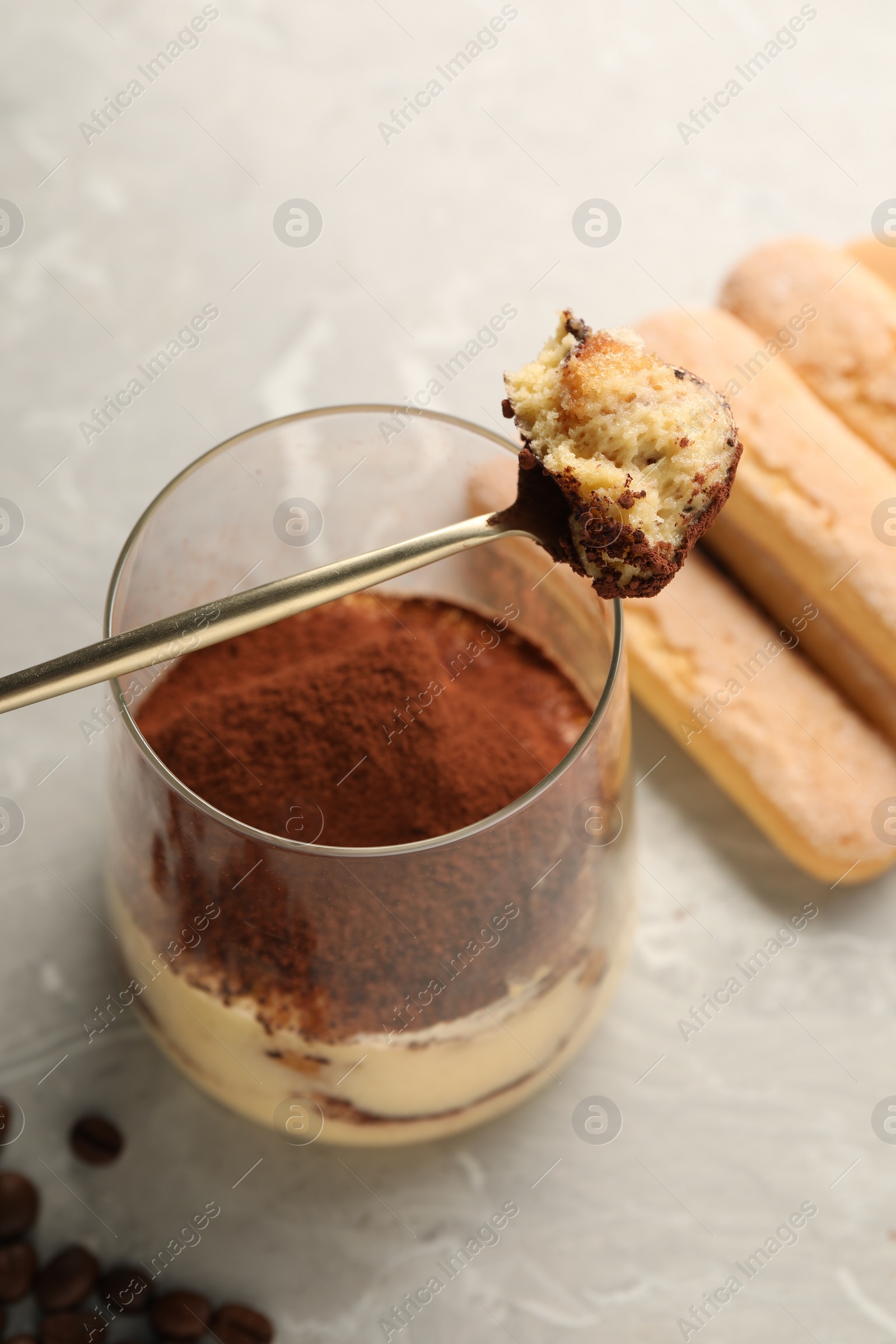 Photo of Tasty tiramisu in glass, coffee beans, spoon and biscuits on light grey table, closeup