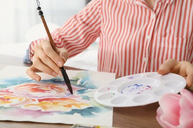 Woman painting flowers with watercolor at wooden table indoors, closeup. Creative artwork