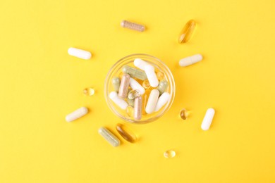 Photo of Different vitamin pills in glass bowl on yellow background, top view