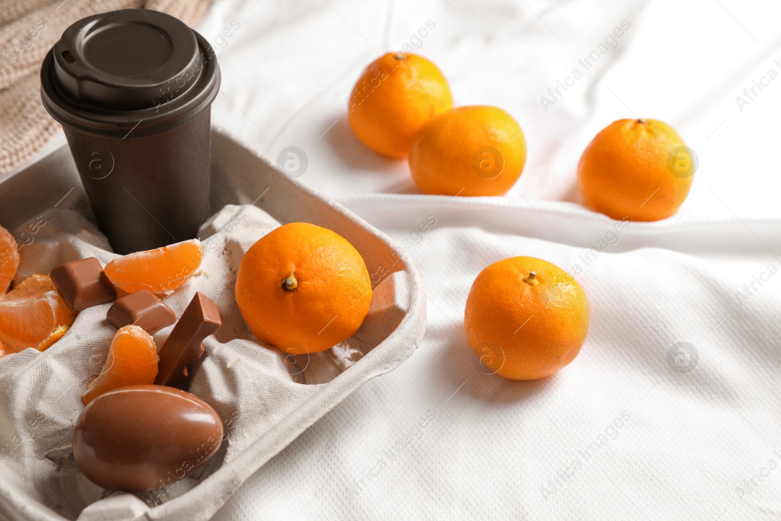 Photo of Delicious ripe tangerines, cup with drink and chocolates on white bedsheet