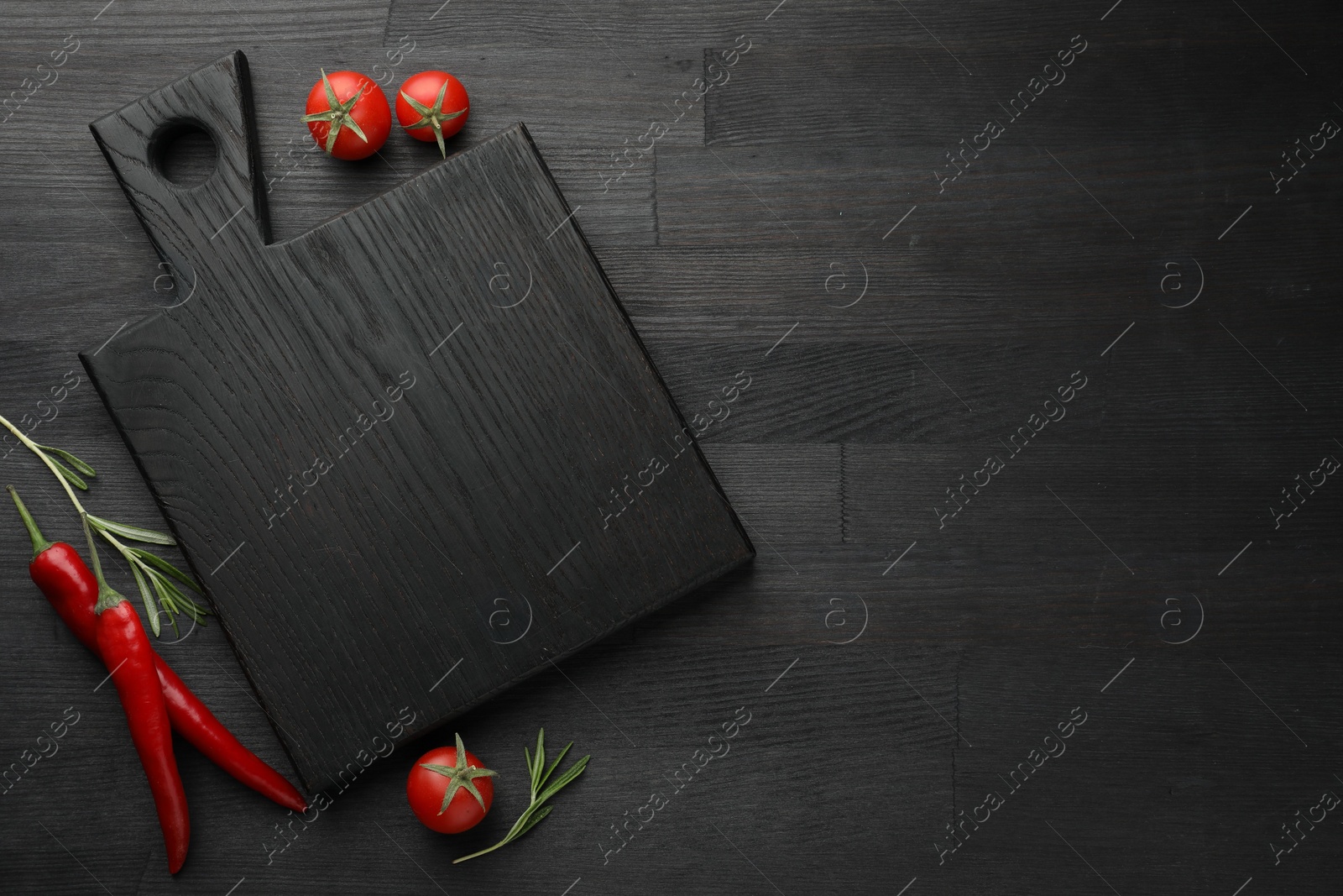 Photo of Cutting board, rosemary, chili peppers and tomatoes on black wooden table, flat lay. Space for text