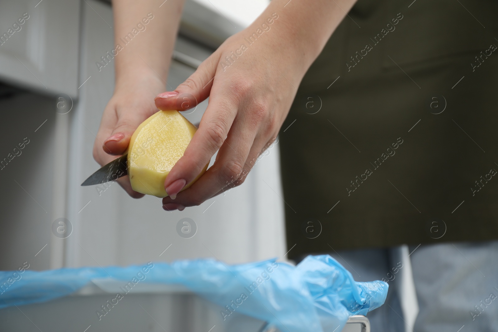 Photo of Woman peeling fresh potato with knife above garbage bin indoors, closeup