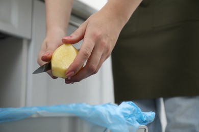 Woman peeling fresh potato with knife above garbage bin indoors, closeup