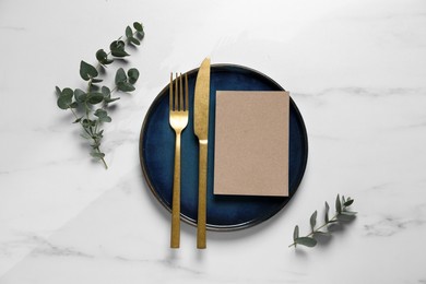 Stylish setting with cutlery, eucalyptus leaves and blank card on white marble table, flat lay. Space for text