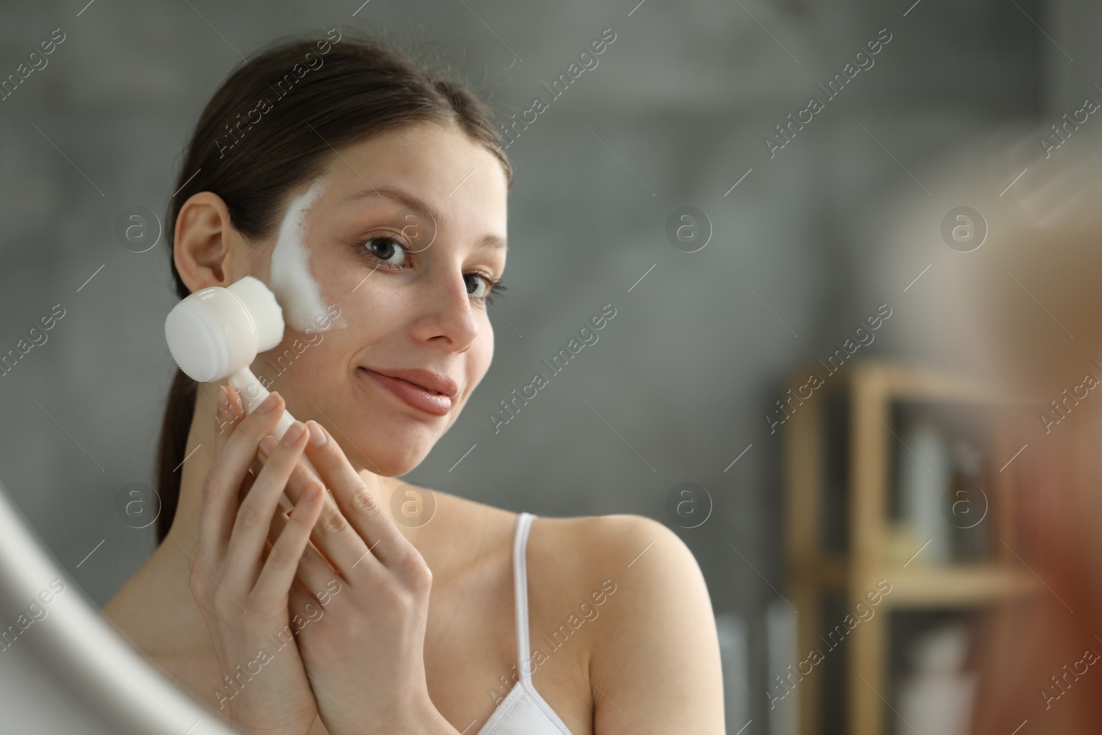 Photo of Young woman washing face with brush and cleansing foam near mirror in bathroom