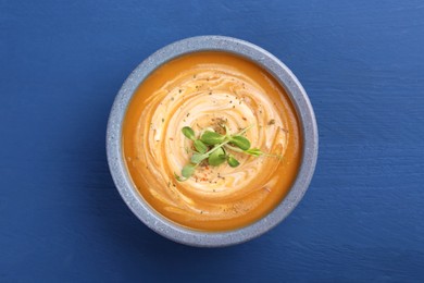 Photo of Bowl of delicious pumpkin soup with microgreens on blue wooden table, top view