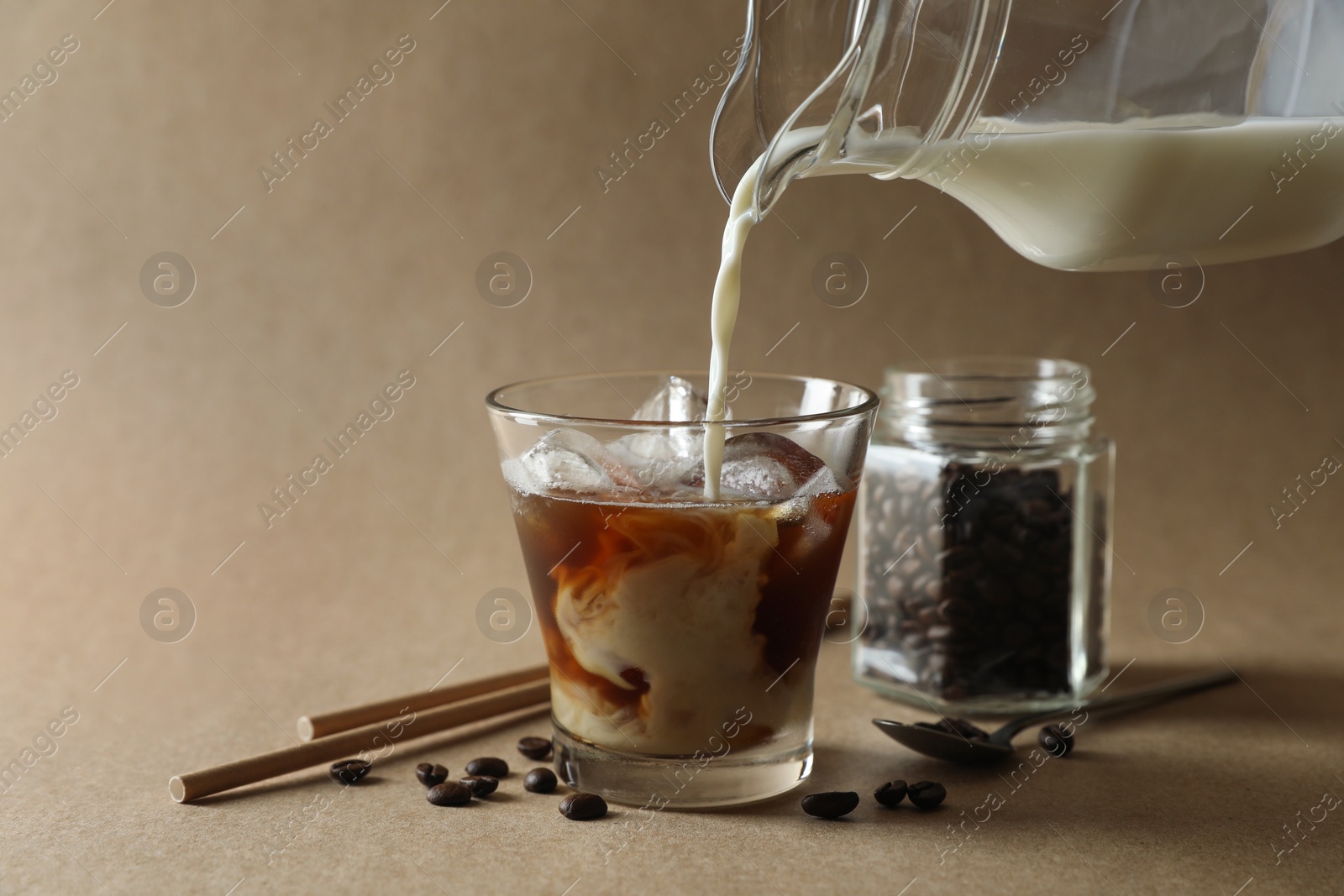 Photo of Pouring milk into glass with refreshing iced coffee on beige background, closeup