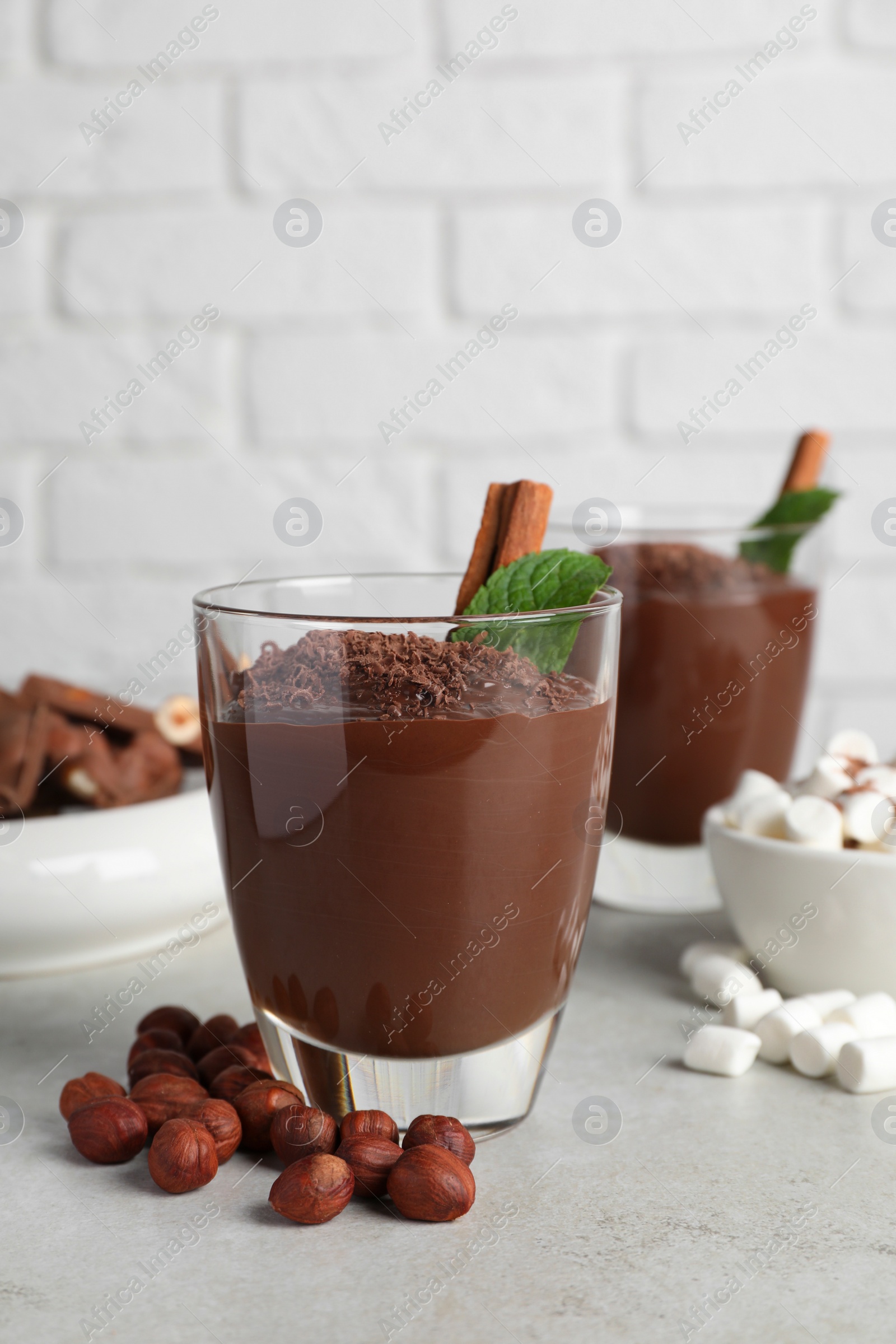 Photo of Cups of delicious hot chocolate with cinnamon stick, mint and hazelnuts on light grey table near brick wall