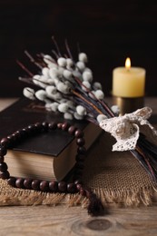 Rosary beads, Bible, burning candle and willow branches on wooden table, closeup