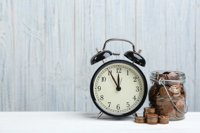 Photo of Black alarm clock, glass jar and coins on white table against wooden background, space for text. Money savings