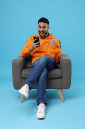 Happy young man using smartphone on armchair against light blue background