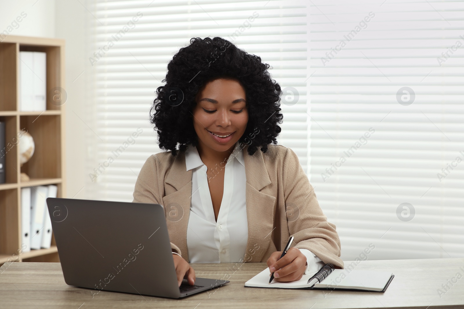 Photo of Happy young woman writing notes while using laptop at wooden desk indoors