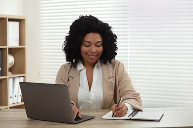 Happy young woman writing notes while using laptop at wooden desk indoors