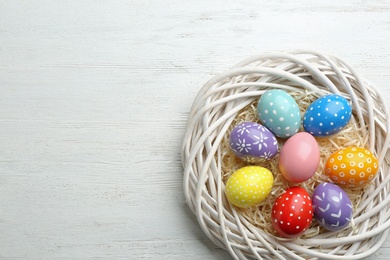 Wicker nest with painted Easter eggs on wooden table, top view. Space for text