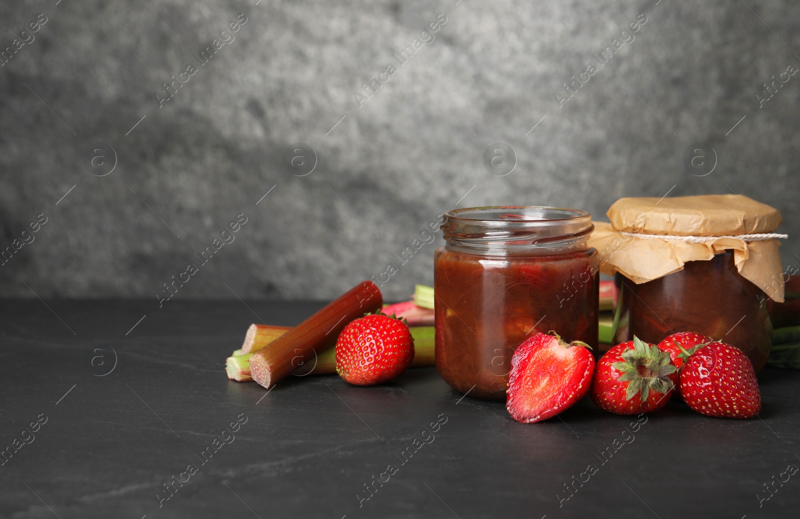 Photo of Jars of tasty rhubarb jam, fresh stems and strawberries on dark textured table. Space for text
