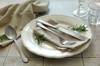 Photo of Stylish setting with cutlery, napkin, rosemary and plate on light tiled table, closeup