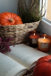 Wicker basket with beautiful heather flowers, pumpkin, burning candles and open book near window indoors
