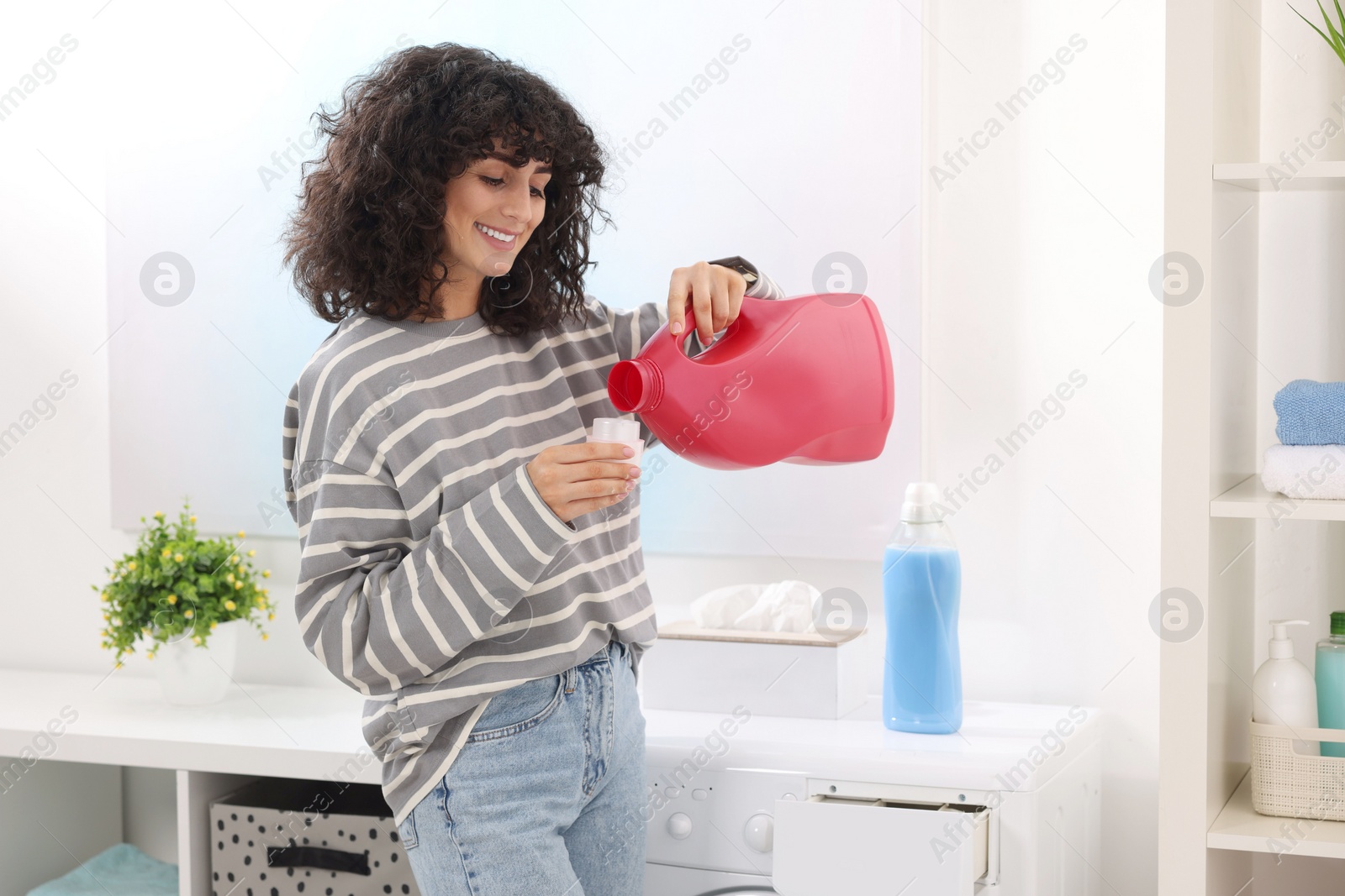 Photo of Happy woman pouring laundry detergent into cap near washing machine indoors