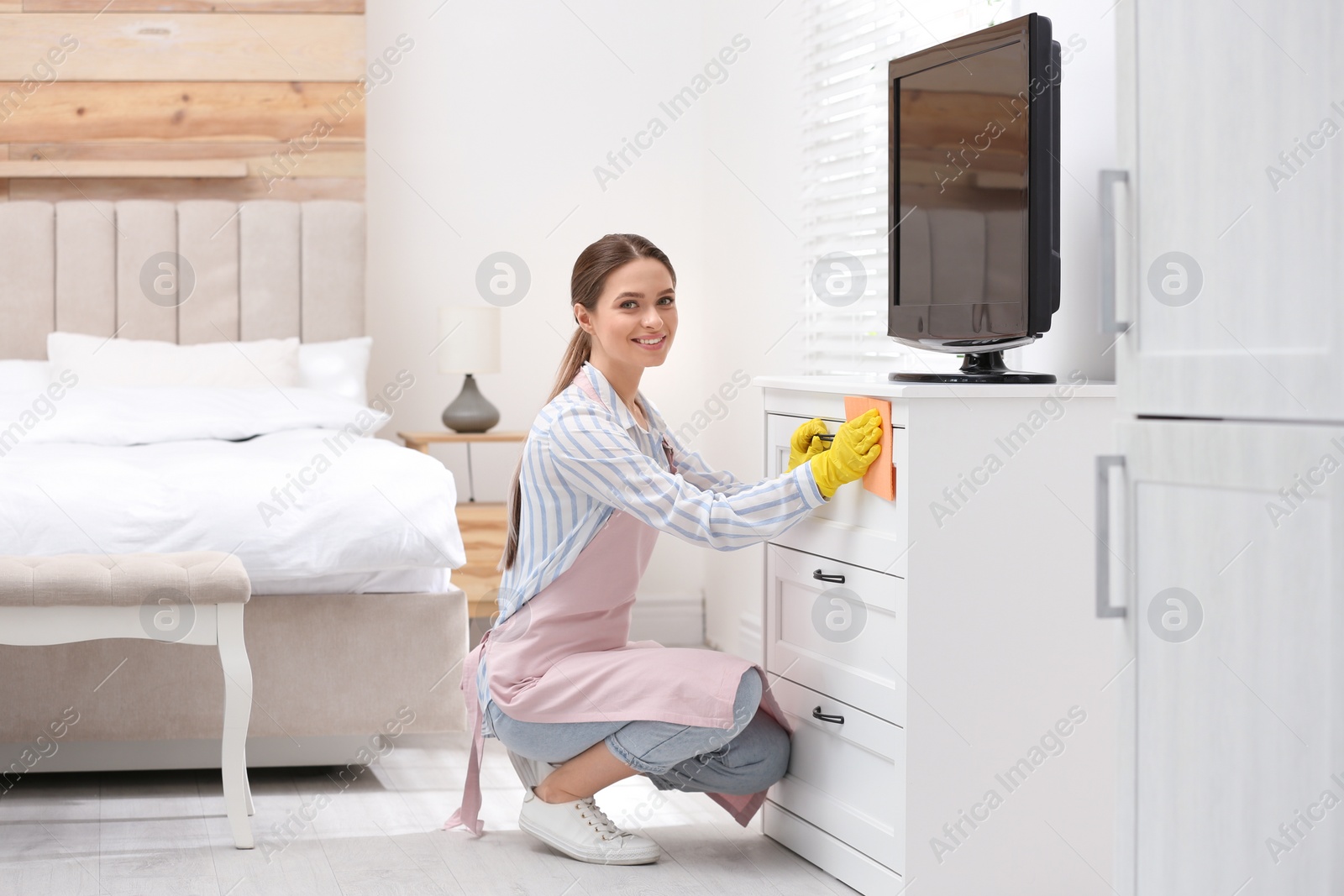 Photo of Young chambermaid wiping dust from furniture in hotel room