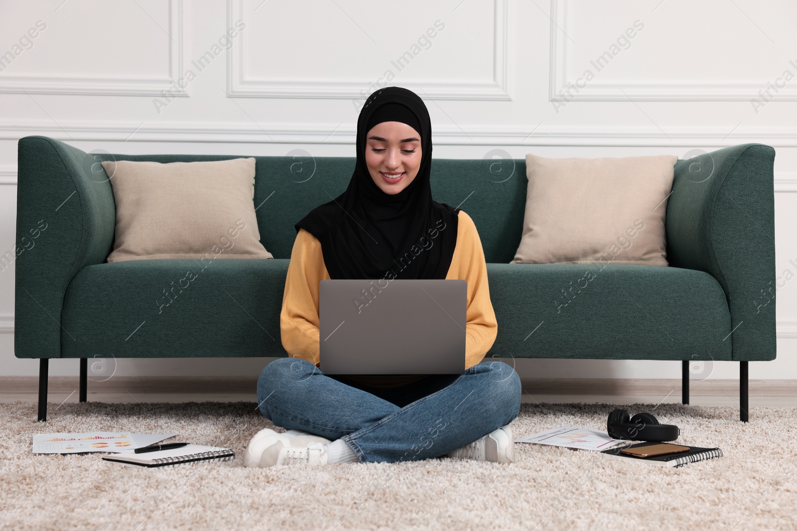 Photo of Muslim woman in hijab using laptop on floor near sofa indoors