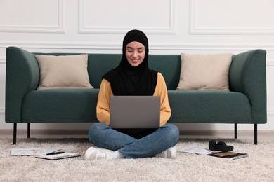 Muslim woman in hijab using laptop on floor near sofa indoors