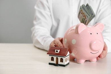 Photo of Man holding piggy bank with dollar banknotes and house model at wooden table, closeup. Space for text. Saving money concept
