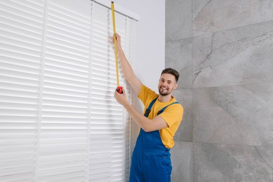 Worker in uniform using measuring tape while installing horizontal window blinds indoors