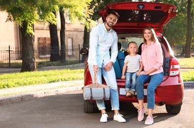 Photo of Happy family with picnic basket sitting in car's trunk on road
