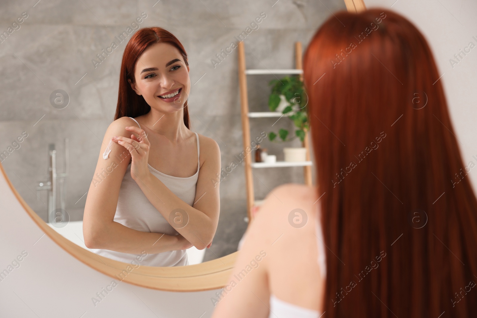 Photo of Beautiful young woman applying body cream onto shoulder near mirror in bathroom