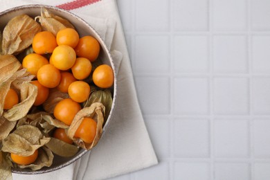 Ripe physalis fruits with calyxes in bowl on white tiled table, top view. Space for text