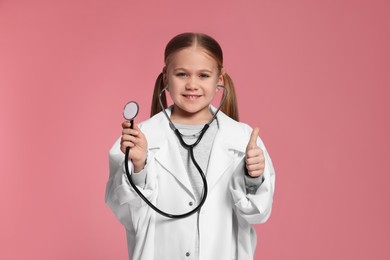 Photo of Little girl in medical uniform with stethoscope showing thumb up on pink background